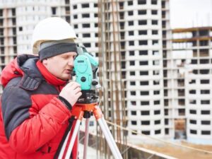 Construction worker in a red jacket and white hard hat uses a surveying instrument on a tripod at a construction site.