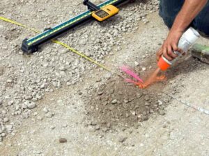 Worker marks a dirt surface with orange spray paint for a digging spot, with a tape measure and level nearby.